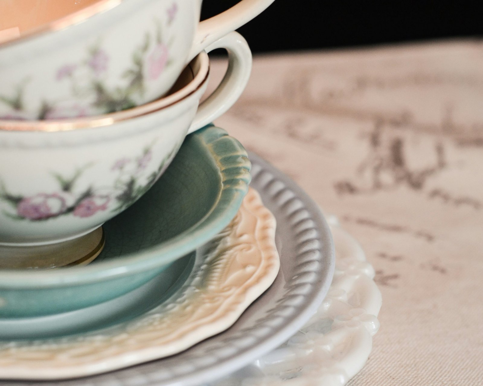 Stacked vintage porcelain teacups and saucers with floral patterns in soft pastel colors, arranged on a table with a blurred handwritten background