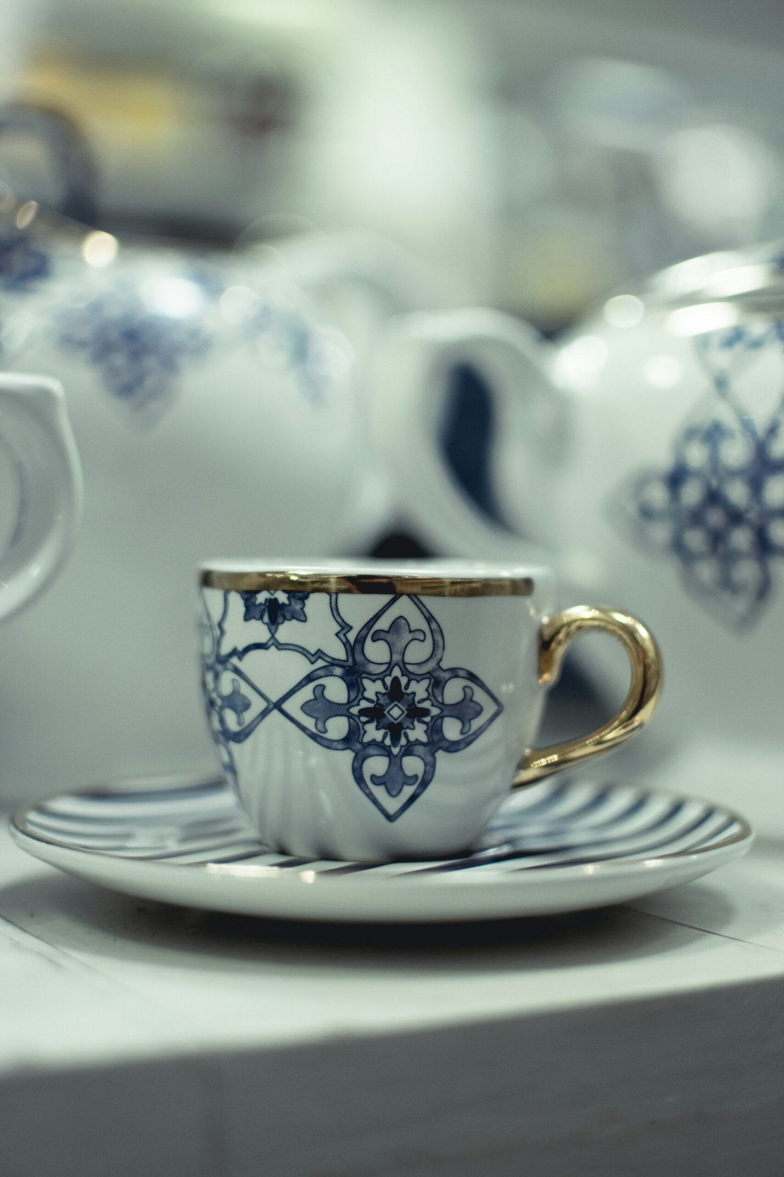 White porcelain espresso cup with blue geometric ornament and gold handle, set on a saucer, with matching cups softly blurred in the background
